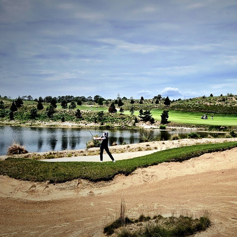 A golfer mid-swing beside a pond and bunker